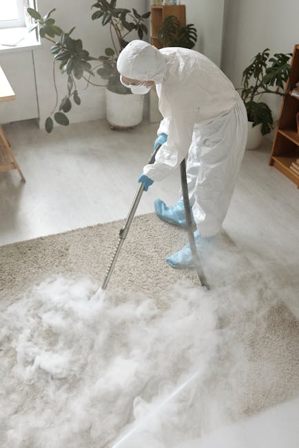 A man in a grey coverall is performing deep cleaning or sanitisation in an auditorium or theatre, using a portable vacuum or extraction machine on the carpeted floor, which appears dark and slightly t